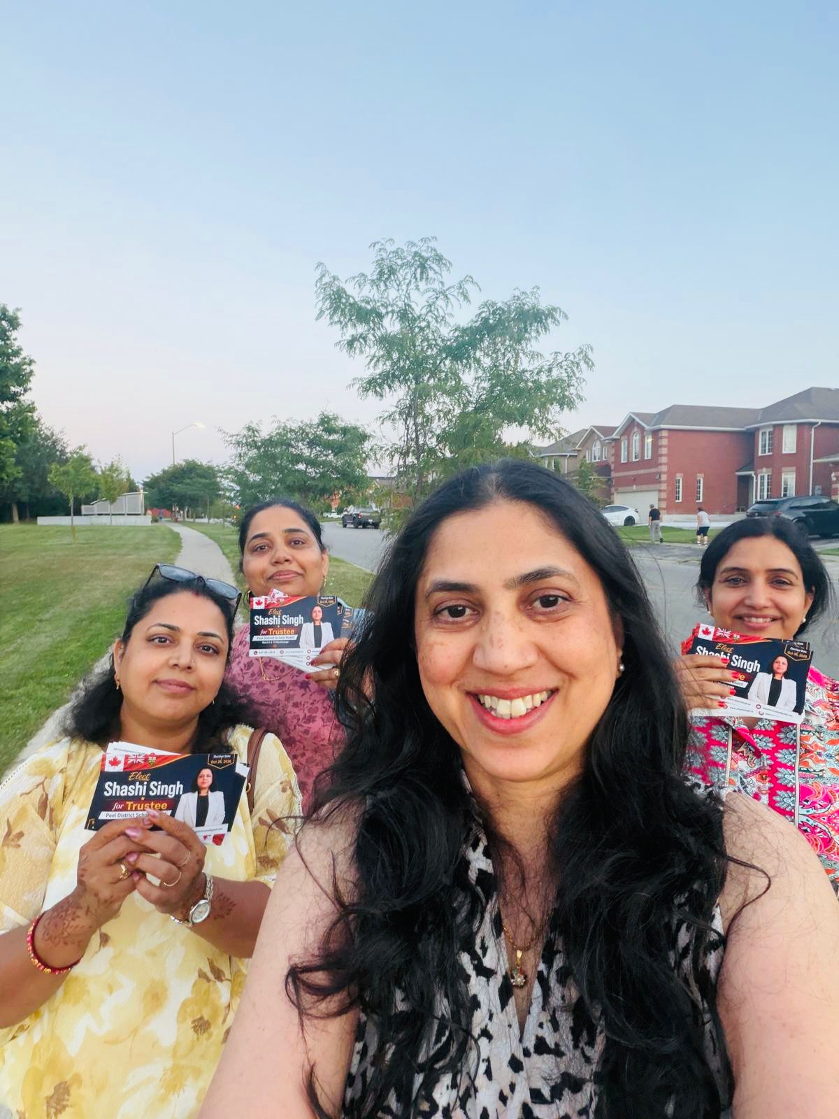 Community supporters smiling and holding Elect Shashi Singh for Trustee flyers in a local park